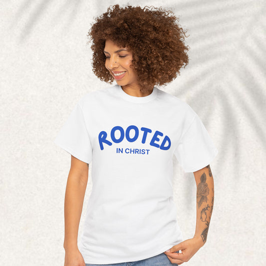 A woman with curly brown hair wearing a white t-shirt with 'ROOTED IN CHRIST' printed in blue on the front, set against a light textured background with soft leaf shadows.